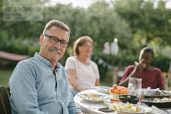 Porträt eines älteren Mannes  der lächelt  während er während einer Gartenparty gegen die Familie am Tisch sitzt