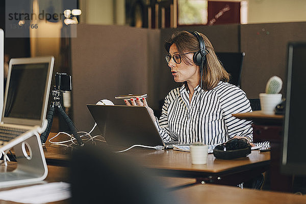 Reife Geschäftsfrau mit Kopfhörern beim Telefonieren mit dem Handy im Büro