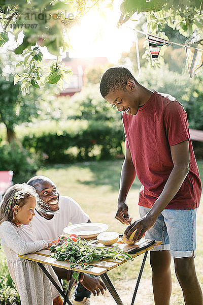Junge schneidet Melone auf dem Tisch  während Vater und Schwester ihn während der Gartenparty anschauen