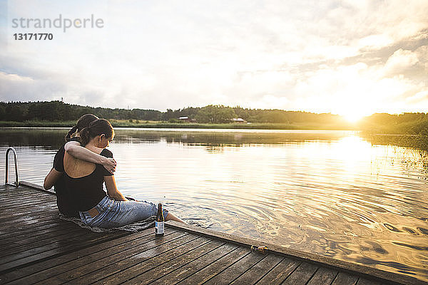 Freundinnen umarmen sich  während sie bei Sonnenuntergang auf dem Steg über dem See gegen den Himmel sitzen