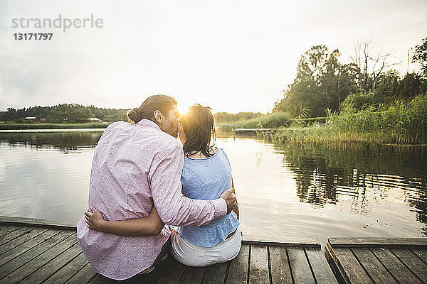 Rückansicht eines Freundes  der seine Freundin küsst und mit umarmten Armen auf dem Steg über dem See bei Sonnenuntergang sitzt