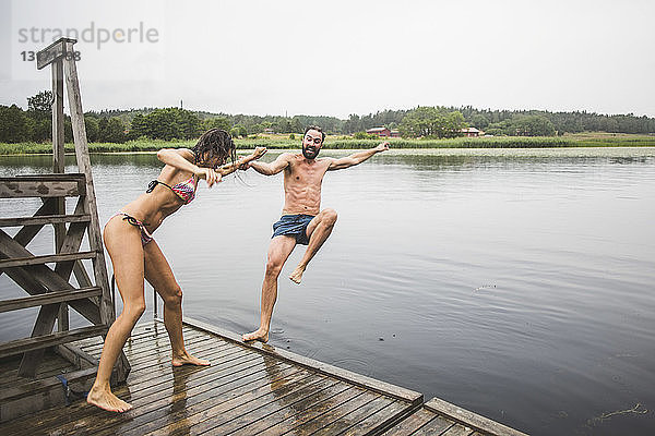 Verspielte Frau in voller Länge schiebt einen männlichen Freund in den See  während sie während eines Wochenendausflugs auf einem Steg steht