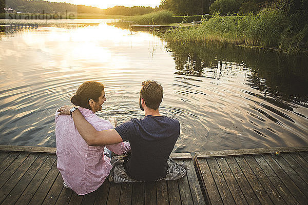 Rückansicht von lächelnden männlichen Freunden  die am Wochenende auf einem Steg über dem See sitzen