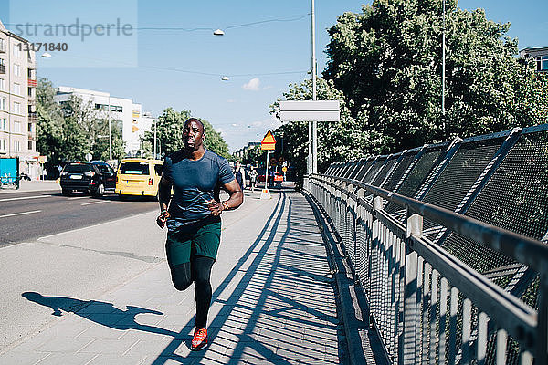 Männlicher Athlet in voller Länge joggt auf dem Bürgersteig an einer Brücke in der Stadt