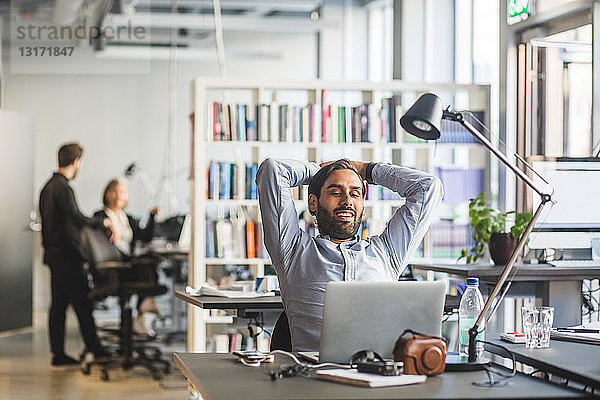 Lächelnder Geschäftsmann schaut auf Laptop  während er im Büro mit Händen hinter dem Kopf sitzt