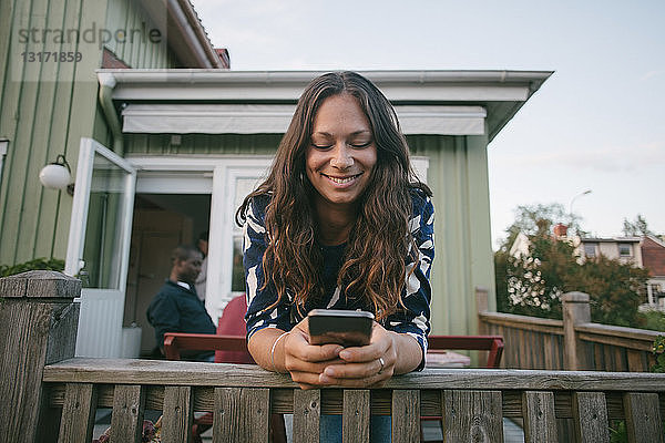Lächelnde mittelgroße erwachsene Frau  die ein Mobiltelefon benutzt  während sie sich auf das Geländer der Veranda lehnt