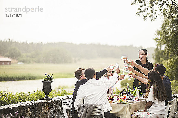 Fröhliche männliche und weibliche Freunde stoßen während der Dinnerparty im Hinterhof auf Weingläser an