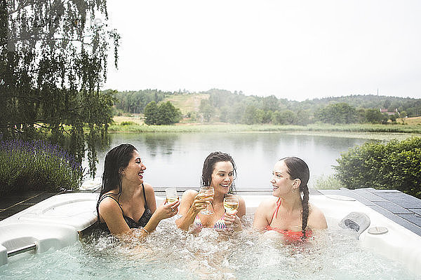Fröhliche Freundinnen genießen Wein im Whirlpool am See während eines Wochenendausflugs