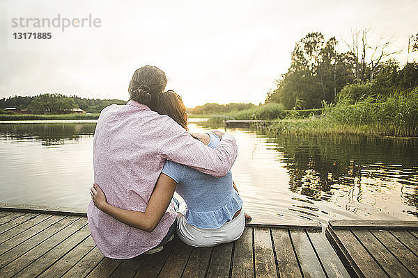 Rückansicht eines Paares  das bei Sonnenuntergang mit umgebundenen Armen auf dem Steg über dem See sitzt