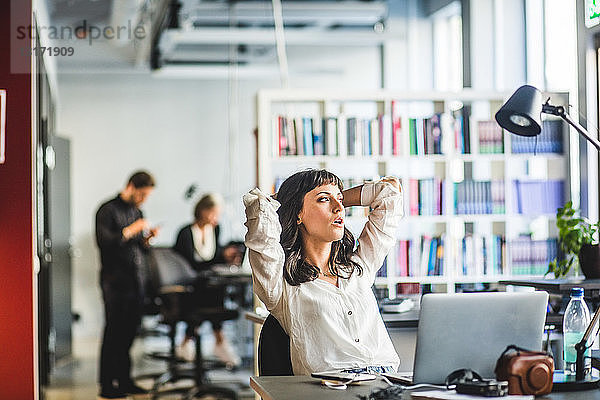 Geschäftsfrau schaut weg  während sie mit Händen hinter dem Kopf am Schreibtisch im Büro sitzt