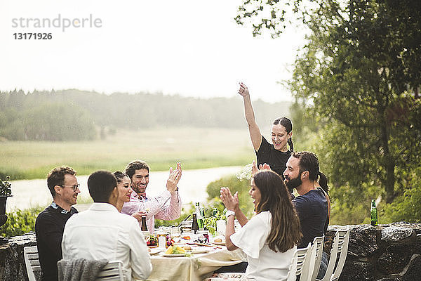 Mittelgroße erwachsene Frau steht mit erhobenem Arm  während Freunde während der Dinnerparty im Hinterhof am Tisch applaudieren
