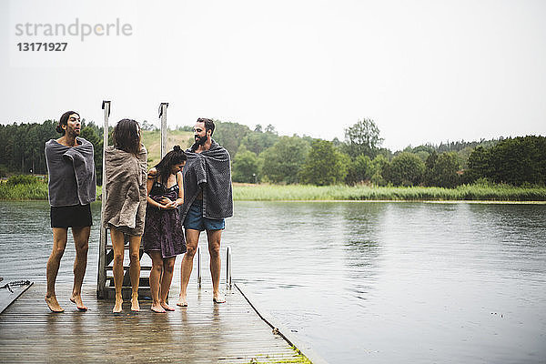 In Handtücher gewickelte Freunde unterhalten sich  während sie auf dem Steg über dem See vor klarem Himmel stehen