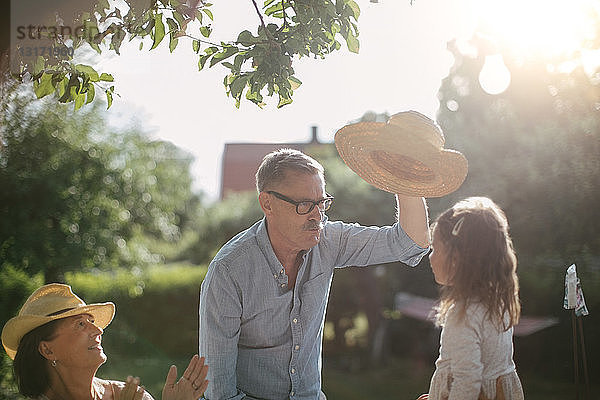 Frau klatscht und schaut älteren Mann an  der an einem sonnigen Tag im Garten einen Strohhut über seine Enkelin hält