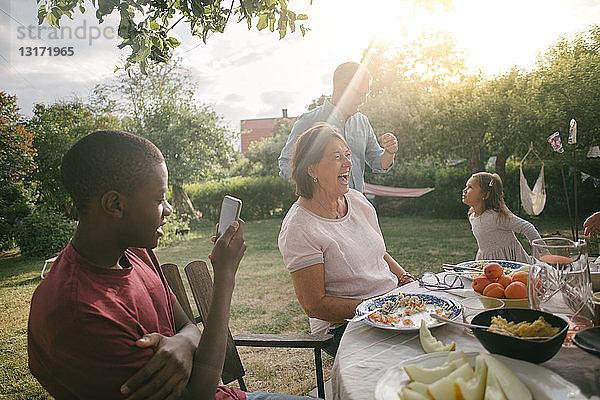 Junge fotografiert fröhliche Familie beim Mittagessen am Tisch während einer Gartenparty