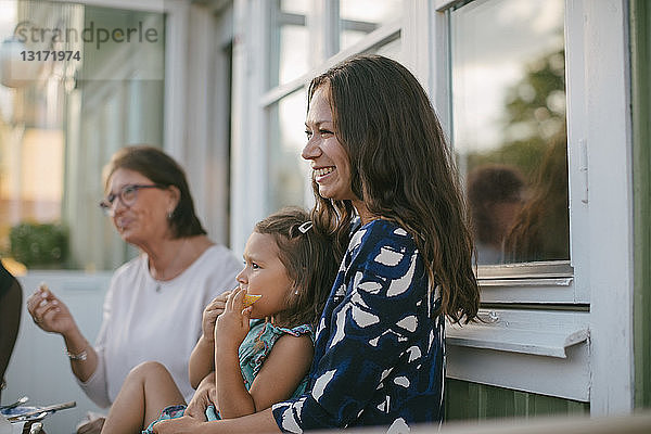 Lächelnde Mutter mit Tochter isst Obst  während sie von einer älteren Frau auf der Veranda sitzt