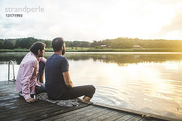Männliche Freunde schauen auf den See  während sie bei Sonnenuntergang auf dem Steg sitzen