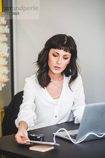 Kreative Geschäftsfrau benutzt Mobiltelefon  während sie im Büro am Schreibtisch sitzt