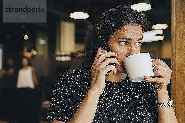 Mittlere erwachsene Geschäftsfrau  die Kaffee trinkt  während sie im Büro ein Smartphone benutzt