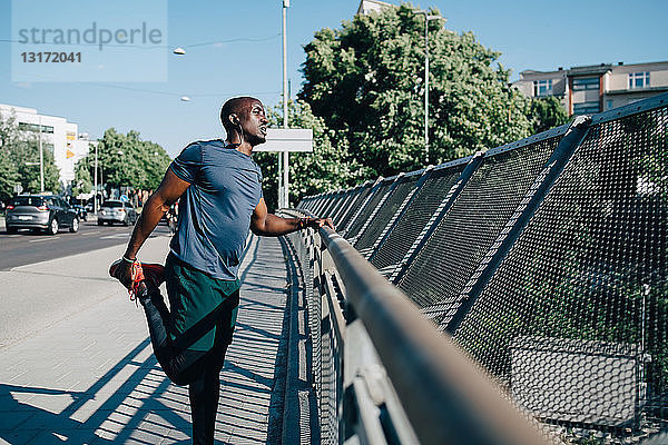 Mittelgroßer erwachsener Sportler  der sein Bein am Geländer einer Brücke in der Stadt streckt