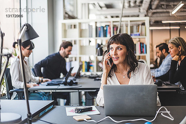 Geschäftsfrau schaut weg  während sie am Schreibtisch im Kreativbüro mit dem Handy telefoniert