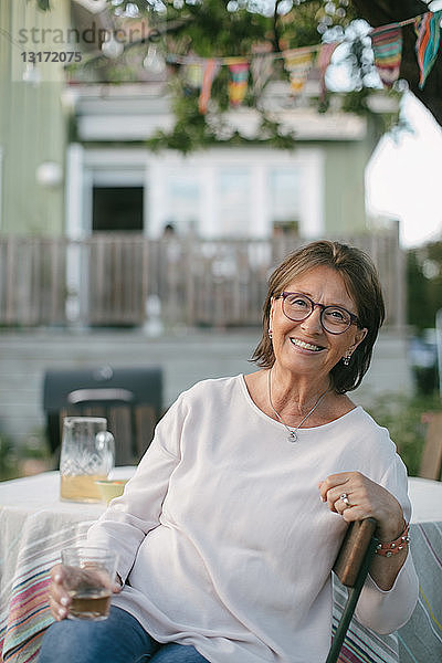Porträt einer lächelnden älteren Frau  die ein Getränk in der Hand hält  während sie auf einem Stuhl am Tisch im Hinterhof sitzt