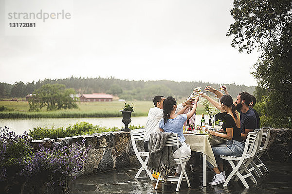 Männliche und weibliche Freunde stoßen am Esstisch auf Getränke an  während sie das Wochenende im Hinterhof verbringen