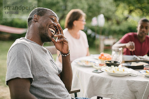 Mittelgroßer erwachsener Mann lächelt  während er während einer Gartenparty am Tisch sitzt