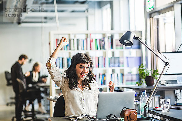 Fröhliche Geschäftsfrau mit geballter Faust schaut im Büro auf Laptop