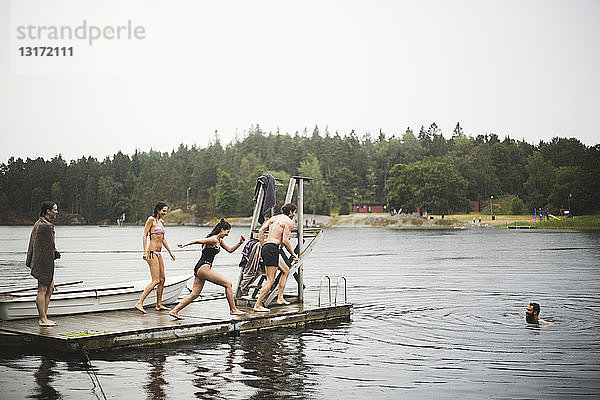 Sorglose männliche und weibliche Freunde rennen auf einem Steg zum Tauchen im See während eines Wochenendausflugs