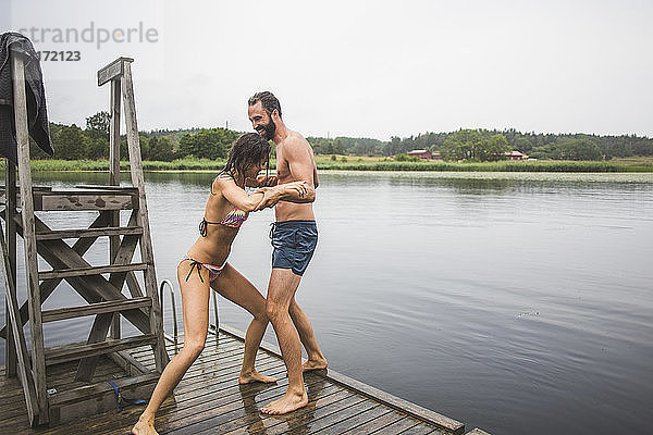 Verspielte Frau schiebt einen männlichen Freund in den See  während sie während eines Wochenendausflugs auf einem Steg steht