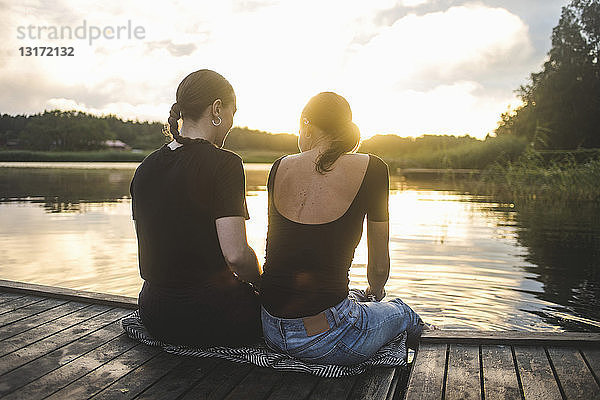 Rückansicht von Freundinnen  die bei Sonnenuntergang auf einem Steg über dem See sitzen