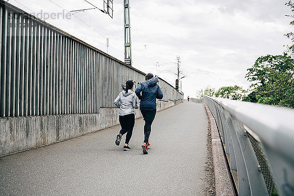 Rückansicht eines männlichen und weiblichen Athleten  die auf einer Fußgängerbrücke in der Stadt joggen