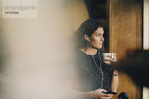 Nachdenkliche Geschäftsfrau  die Kaffee trinkt  während sie im Büro ein Mobiltelefon benutzt
