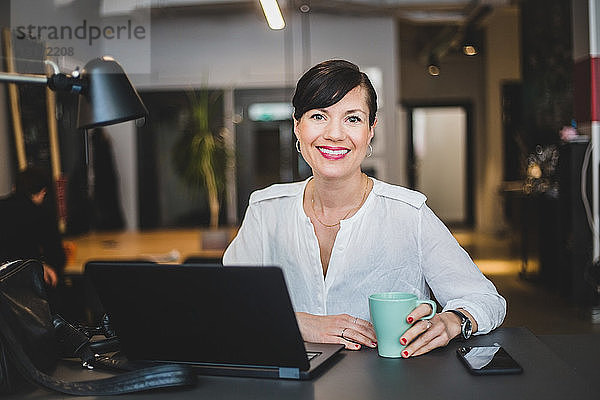 Porträt einer selbstbewussten Geschäftsfrau  die im Büro am Schreibtisch sitzt und einen Kaffeebecher hält