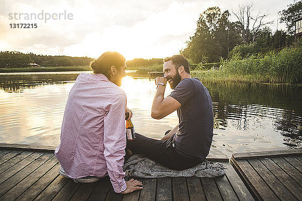 Lächelnde männliche Freunde unterhalten sich  während sie bei Sonnenuntergang auf dem Steg über dem See sitzen