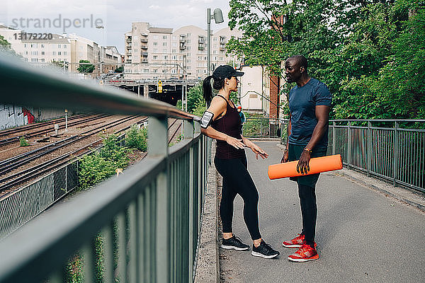 Sportler hält Yogamatte während eines Gesprächs mit einer Sportlerin auf einer Fußgängerbrücke in der Stadt