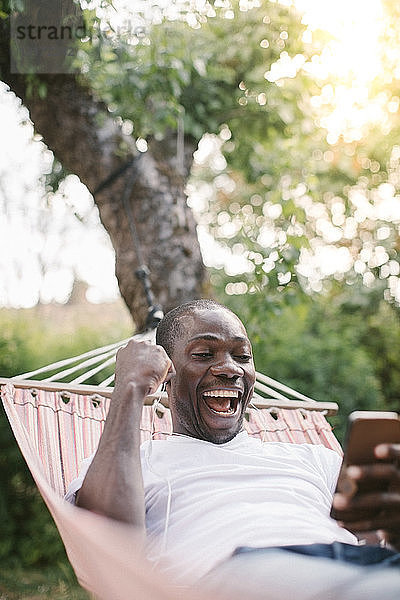 Fröhlicher mittelgroßer Mann schaut auf Handy  während er sich auf einer Hängematte im Hinterhof entspannt