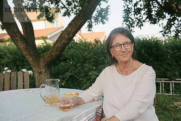 Porträt einer lächelnden Frau  die ein Getränk in der Hand hält  während sie im Hinterhof am Tisch sitzt