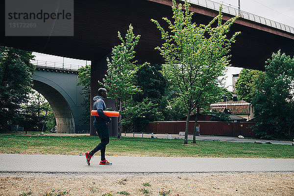 Seitenansicht eines männlichen Athleten  der eine Yogamatte hält  während er auf einem Fußweg in der Stadt läuft