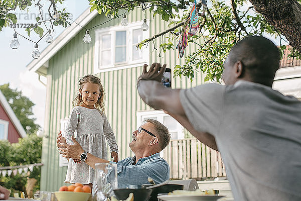 Mann fotografiert Tochter  die während einer Gartenparty am Tisch neben einem älteren Mann steht