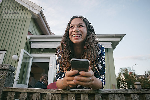 Glückliche Frau im mittleren Erwachsenenalter benutzt Mobiltelefon  während sie sich auf der Veranda an ein Geländer lehnt