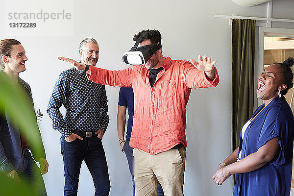 Fröhliche Geschäftsleute betrachten männliche Kollegen  die einen Virtual-Reality-Simulator im Büro benutzen