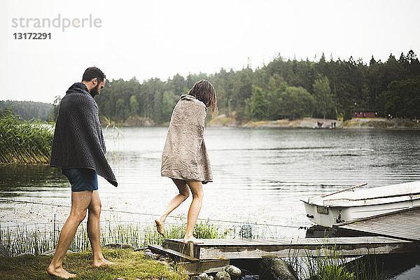 In Handtücher gewickelte Freundinnen und Freunde gehen während eines Wochenendausflugs zum Steg über dem See