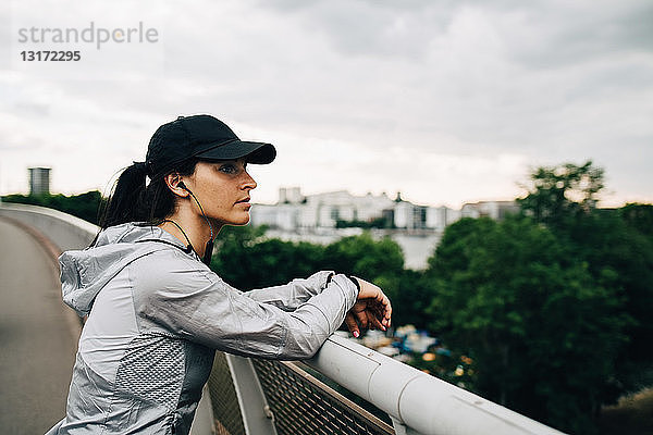 Müde Sportlerin schaut weg  während sie sich am Geländer einer Fußgängerbrücke in der Stadt anlehnt