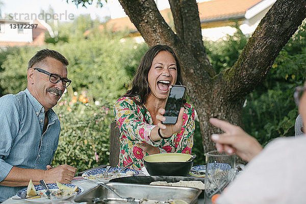 Fröhliche Frau zeigt einer älteren Frau ein Smartphone  während sie während einer Gartenparty am Tisch sitzt
