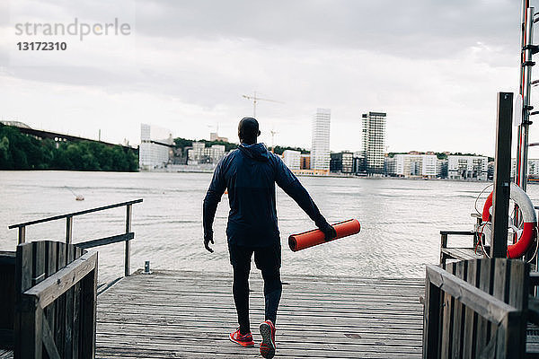 Rückansicht eines männlichen Athleten  der eine Yogamatte in der Hand hält  während er in der Stadt auf dem Pier über das Meer geht