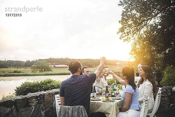 Männliche und weibliche Freunde stoßen bei Sonnenuntergang am Esstisch auf Getränke gegen den Himmel an