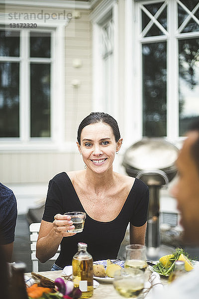 Porträt einer lächelnden Frau mittleren Alters  die ein Getränk im Glas hält  während sie am Esstisch vor der Villa sitzt