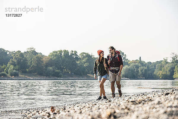 Lächelndes junges Paar mit Rucksäcken beim Spaziergang am Flussufer