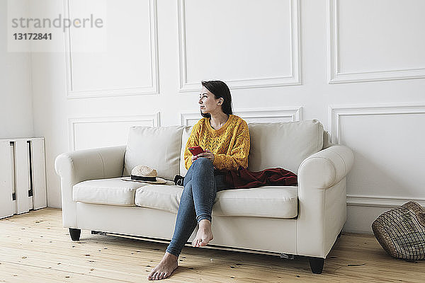 Entspannte junge Frau mit Smartphone sitzt im neuen Zuhause auf der Couch und schaut in die Ferne